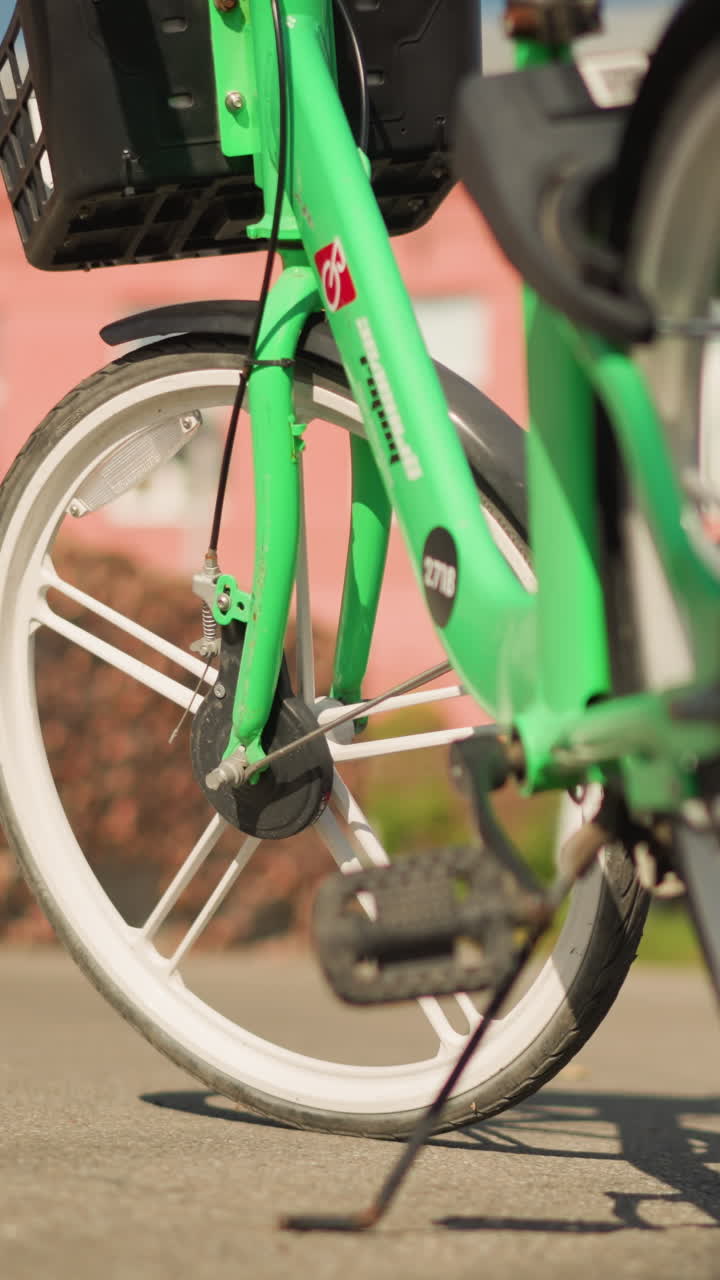 킥스탠드와 그림자가 드리워진 포장 도로 위에 녹색 자전거 바퀴가 클로즈업되어 있고, 밝은 햇빛이 따뜻한 색조를 드리우며, 흰색 림과 스포크, 질감이 있는 아스팔트, 흐릿하게 보이는 주거용 건물 배경, 도시의 임대 자전거가 보이네요.