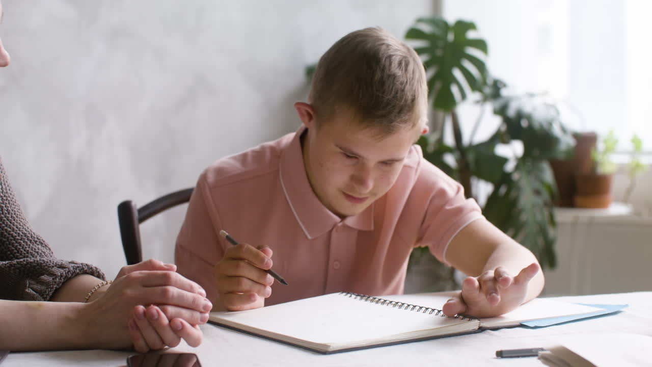 Close-up view of a boy with down syndrome doing homeworks sitting at table in the living room at home. His mother helps him