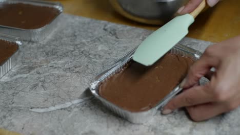 pasta de chocolate esparcida sobre un brownie en un recipiente de aluminio usando una cuchara de hornear, filmada de cerca en estilo cámara lenta