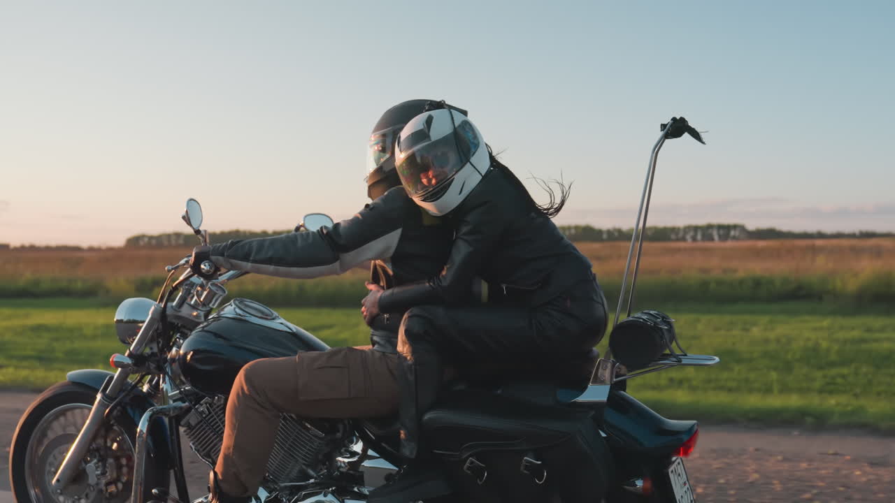 Side view of couple wearing helmets riding motorcycle during scenic road trip, passenger embracing rider as they travel through open countryside with green fields and trees