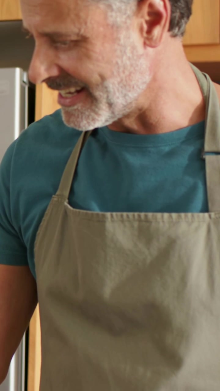 Vídeo vertical: Homem de avental verde-oliva preparando uma refeição na bancada da cozinha, olhando para baixo, com pulseira visível.