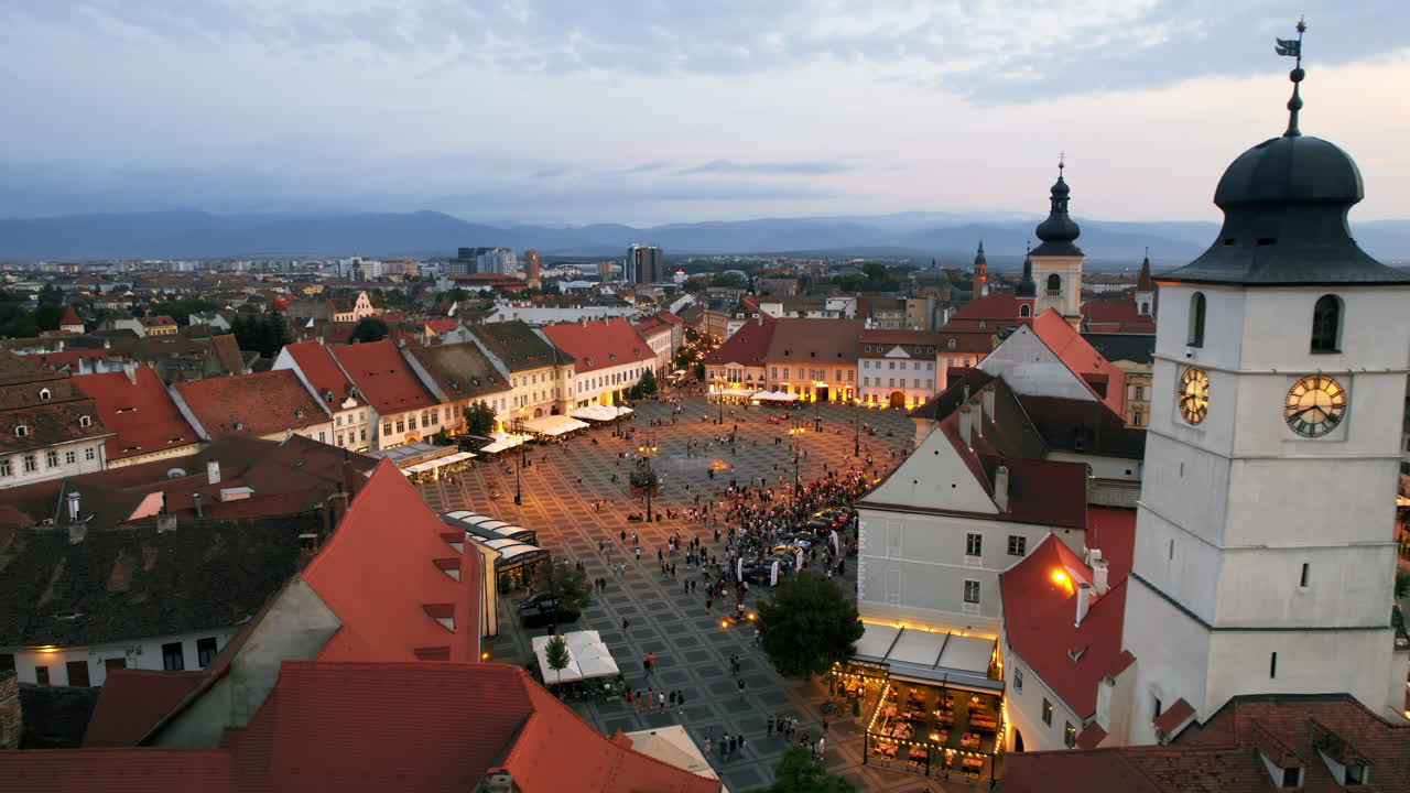 Lotniczy widok z drona na Stare Miasto w Sybinie o zachodzie słońca, Rumunia. Wielki Rynek z Wieżą Rady, stare budynki dookoła, wąskie uliczki, ludzie, oświetlenie