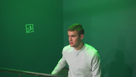 gentleman in shirt and trousers walking up stairs, holding rail with serious expression under green lighting. shadows cast on walls create a moody atmosphere as he moves towards a billiard room