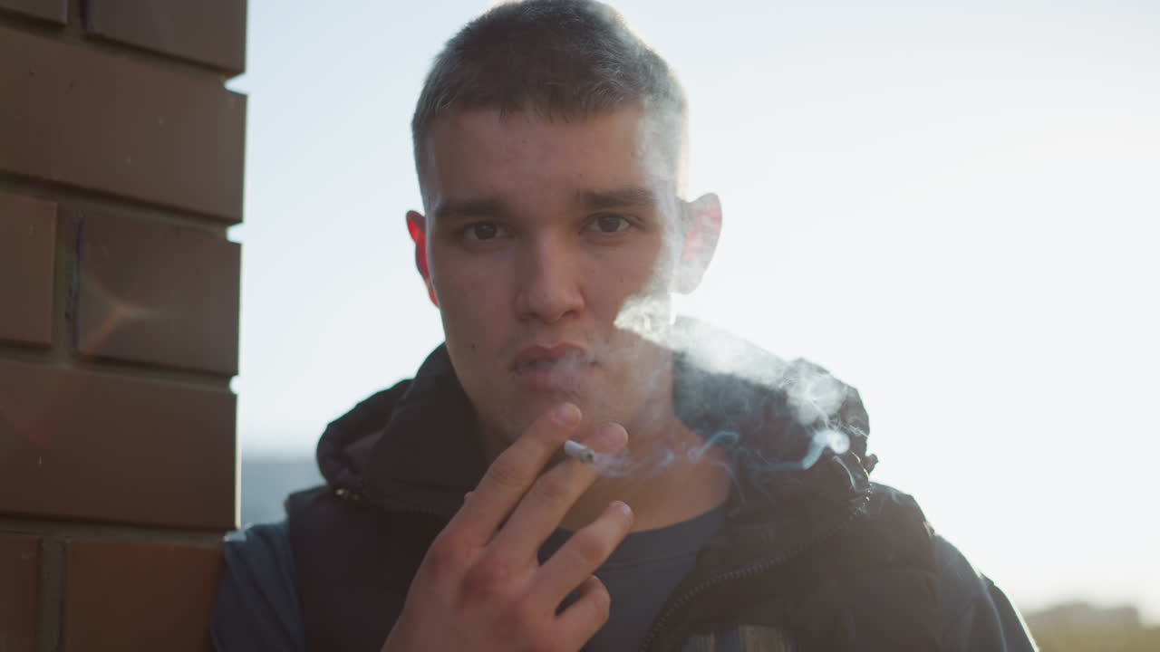 young man stands near brick wall holding cigarette close to mouth, puffing smoke while staring forward with thoughtful expression, soft sunlight creating dramatic backlight effect around smoke