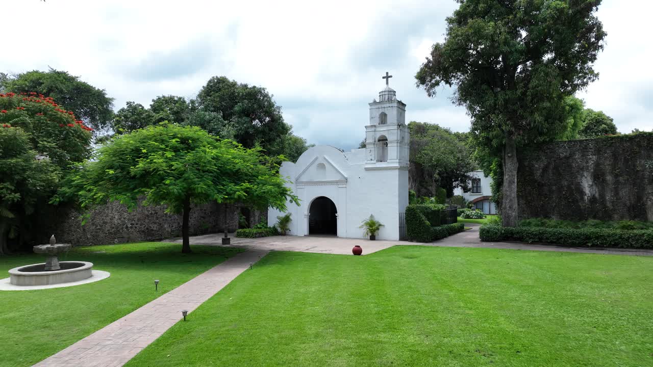 Exterior view of a white colonial church with a cross on top, surrounded by lush greenery
