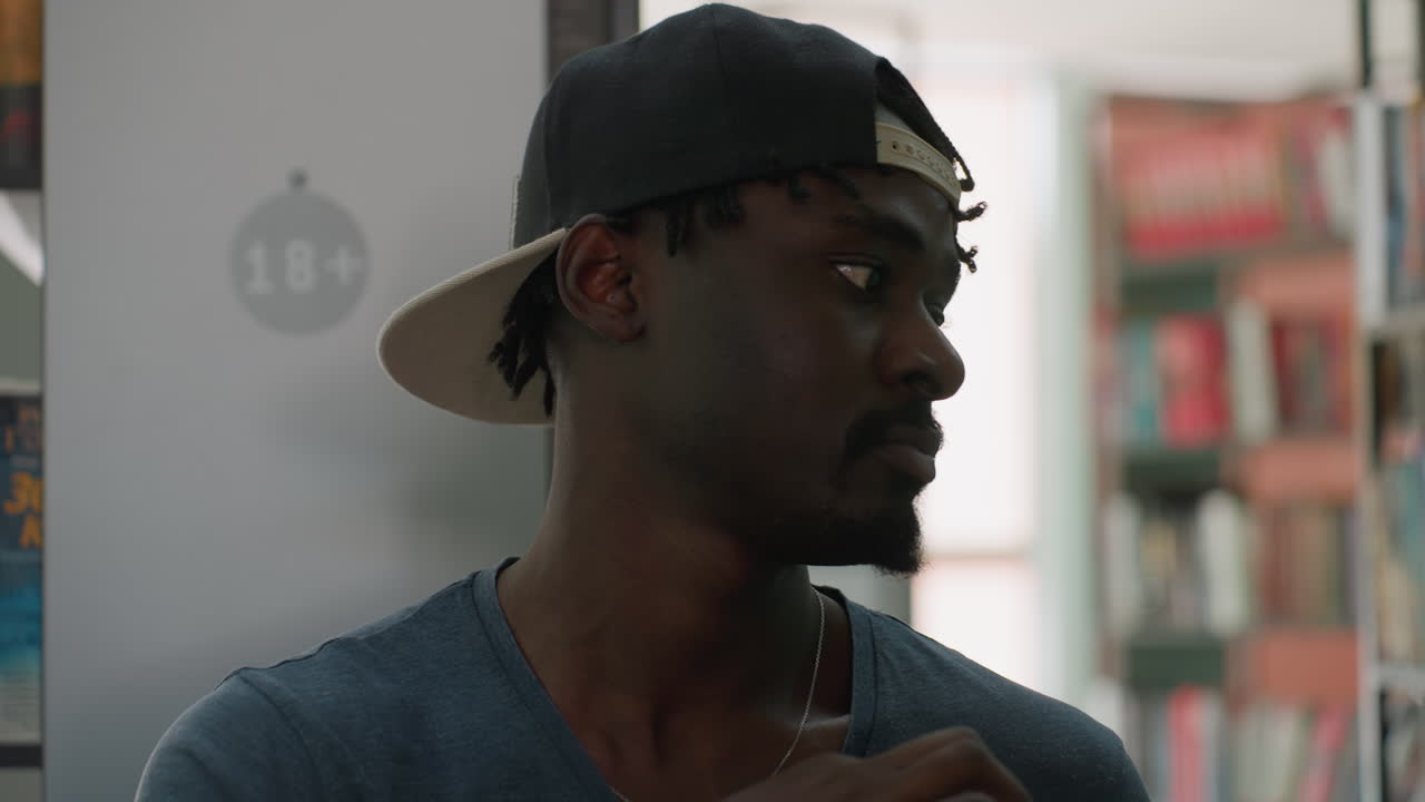 Young man wearing cap holding stack of books in hands while standing in library aisle, glancing around with thoughtful expression, surrounded by colorful bookshelves