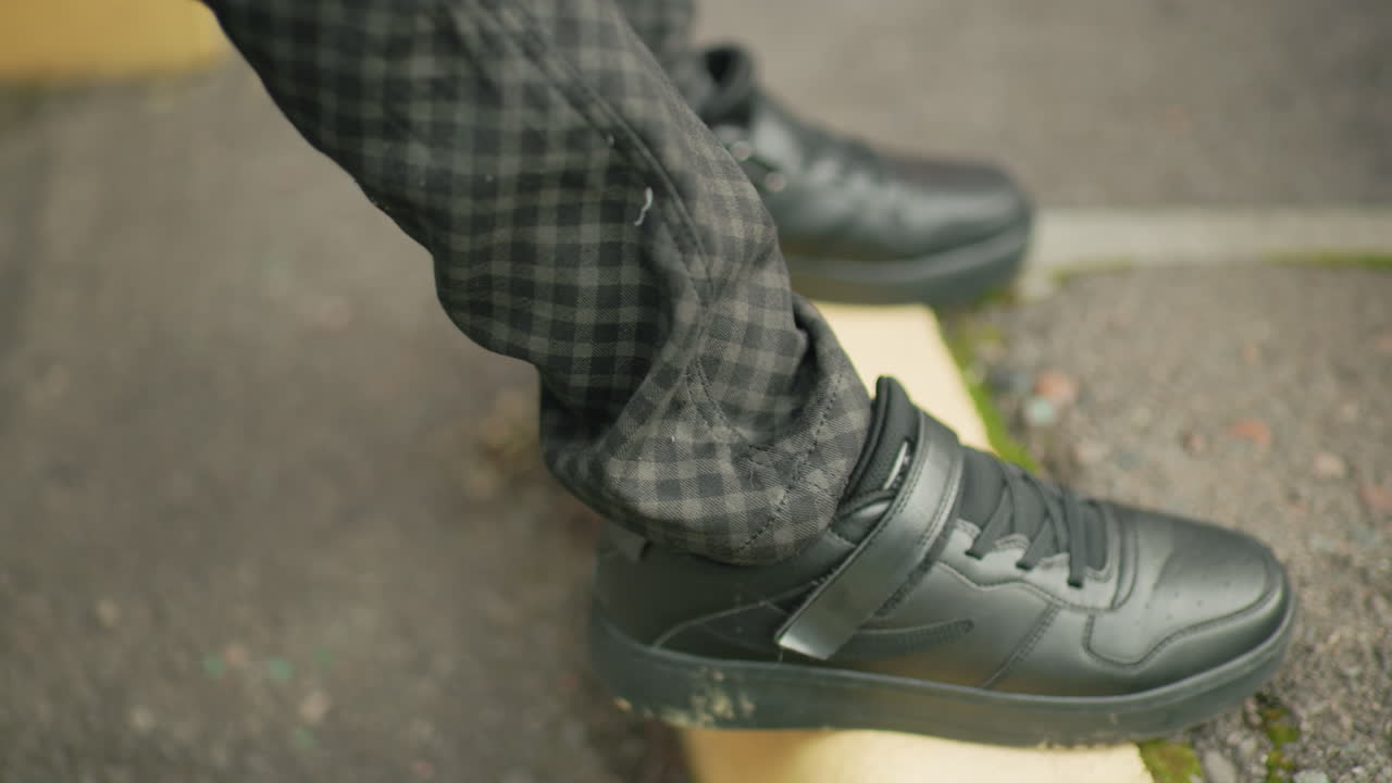 Close up of person legs wearing black sneakers and patterned pants, foot stepping onto yellow pavement curb outdoors, highlighting footwear detail, movement in an urban setting