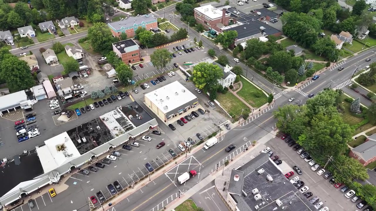 Luftfoto med drone ovenfra som flyr over en liten amerikansk by, og viser sentrumsområdet med gater, trafikk, bygninger, parkeringsplasser og frodige grønne trær på en skyet sommerdag.
