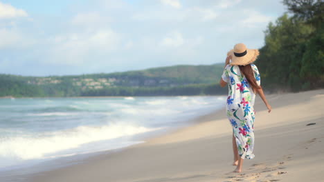 A-woman-in-a-colorful-print-coverup-and-straw-sun-hat-walks-along-a-windswept-beach-as-big-waves-crash-along-the-shore