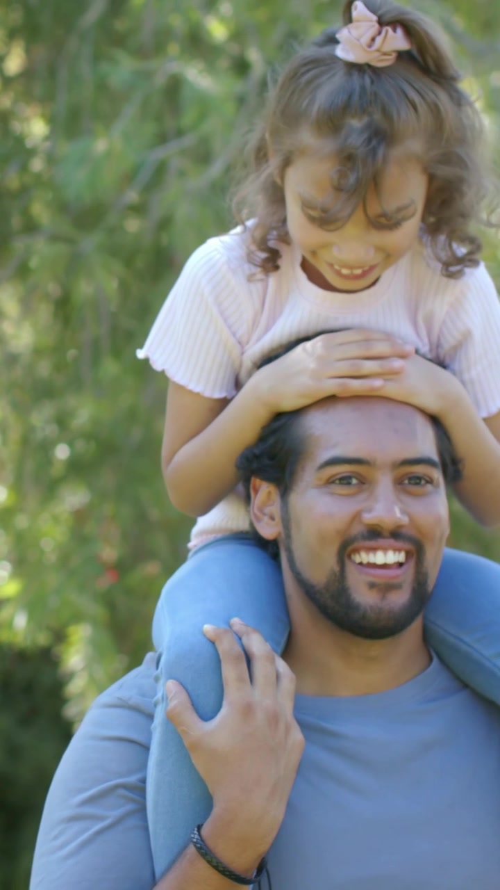 Video vertical: Papá cargando a la niña firmemente sobre sus hombros en el jardín, mamá alcanzando una goma para el pelo.
