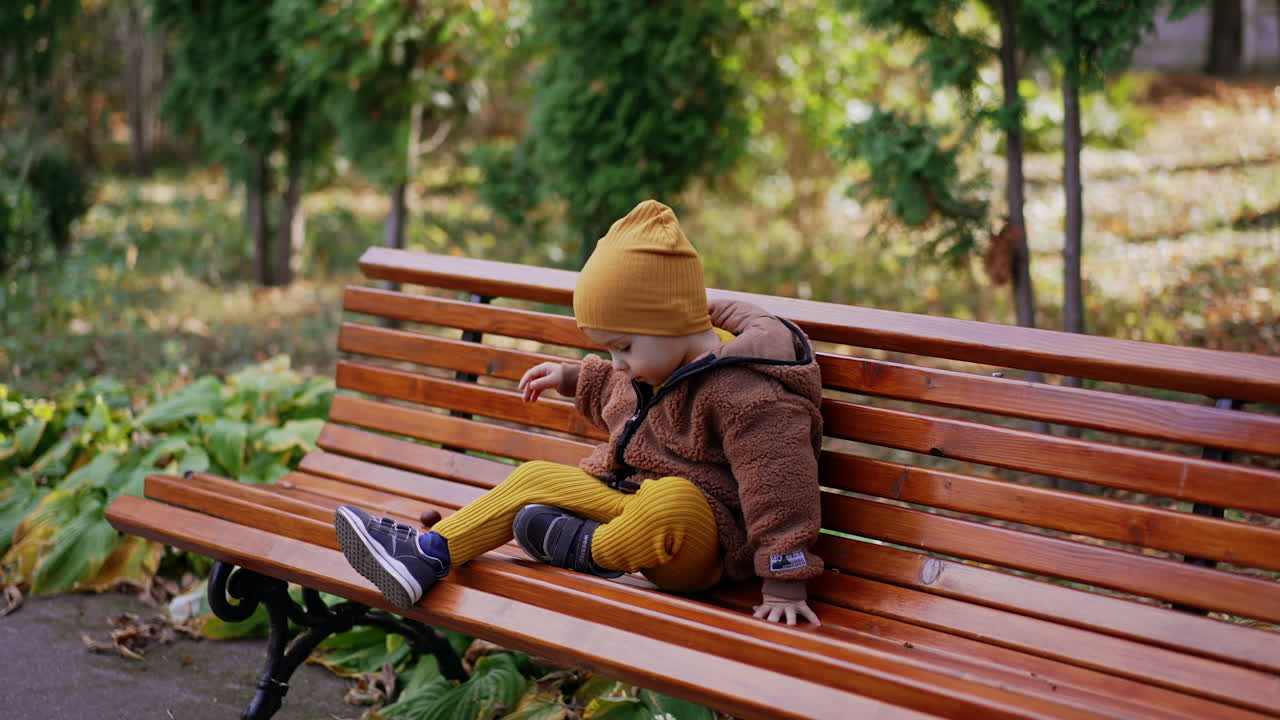 Caucasian toddler crawls by the wooden bench and sits comfortably. Baby boy having rest in the park in autumn.