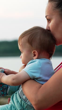 mother is holding a baby in her arms sitting near the river in summer evening. loving mommy kissing her son in head. blurred backdrop. vertical video