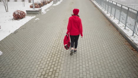 back view of athlete wearing red winter jacket and head warmer holding backpack in her left hand, walking along snowy fog-covered path surrounded by benches and trees dusted with snow