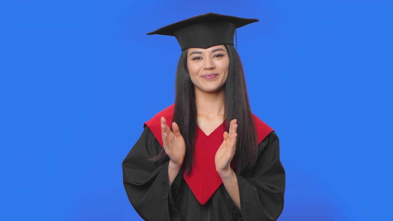 Portrait of female student in cap and gown graduation costume clapping happily. Young brunette woman posing in studio with blue screen background. Close up. Slow motion ready 59.94fps