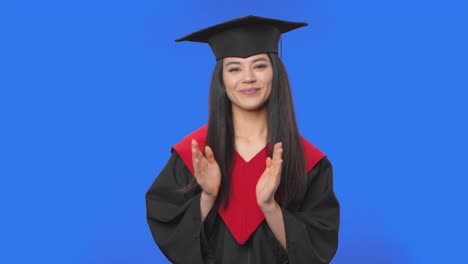 retrato de una estudiante en traje de graduación de gorra y túnica aplaudiendo felizmente. joven mujer morena posando en el estudio con fondo de pantalla azul. primer plano. cámara lenta lista 59.94fps