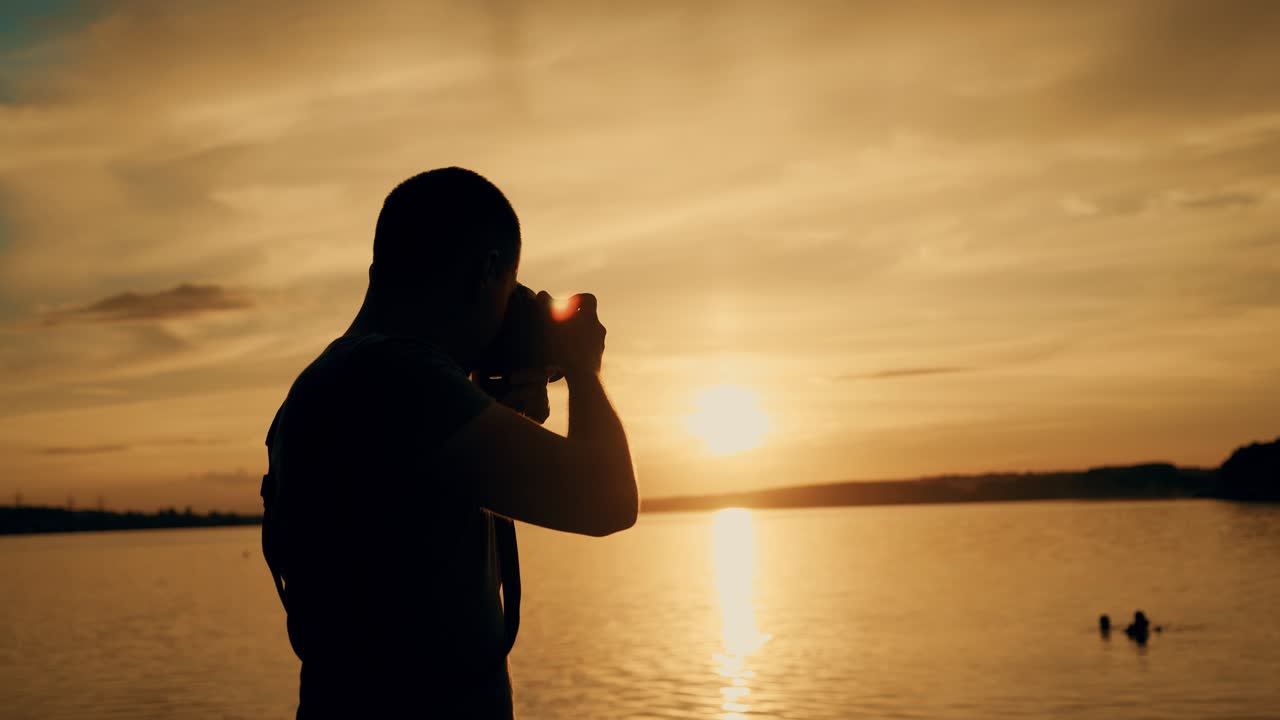 A man with a camera in his hands is photographing the sunset by the river on the background of people bathing. Close-up