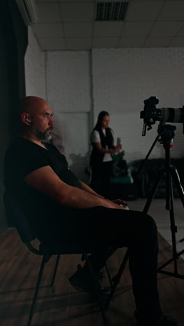 calm male photographer sits in studio looking at camera. girl at backdrop takes video on her phone. backstage of blog footage. vertical video