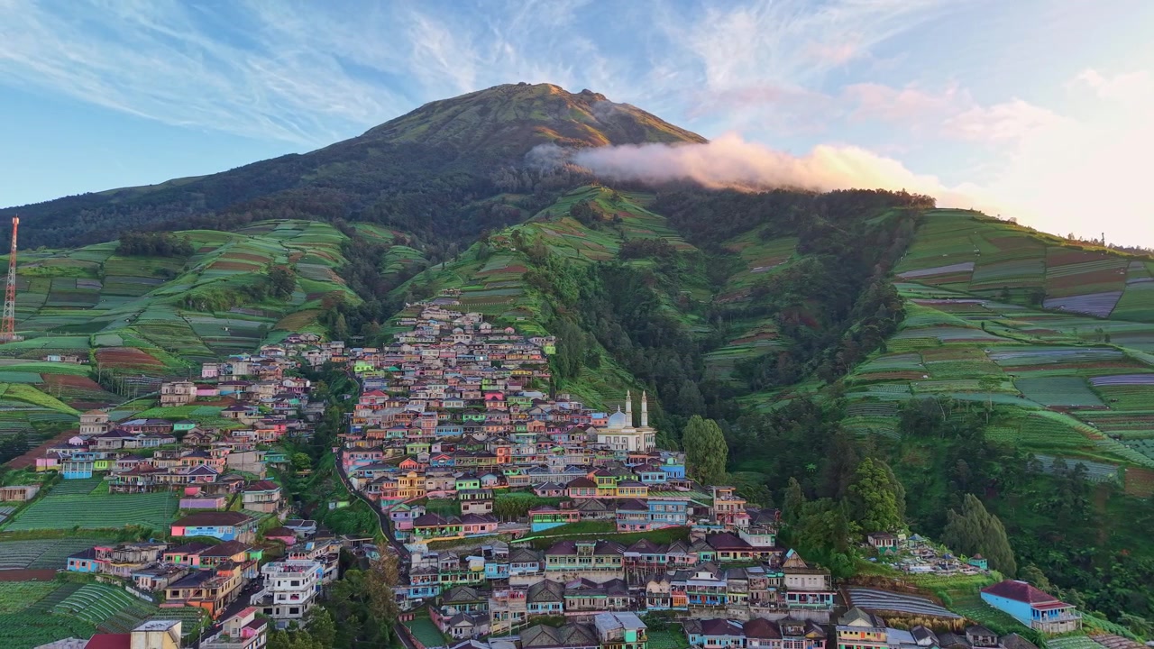 Vista panoramica con drone di un villaggio agricolo in montagna con campi verdi e rigogliosi e un'imponente vetta montuosa. Paesaggio naturale perfetto per contenuti turistici.