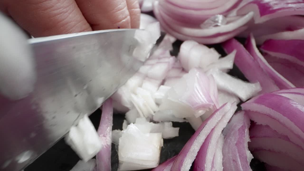 A chef is carefully chopping a purple onion on a black slate cutting board