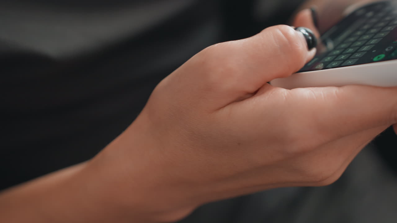 Closeup female hands texting on smartphone, black nail polish, gentle thumb motions, dim indoor lighting, shallow depth of field, blurred couch background, modern keyboard visible, intimate personal