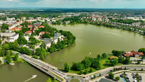 Aerial-view-of-a-lakeside-Iława-town-with-vibrant-buildings,-green-trees,-and-a-large-body-of-water