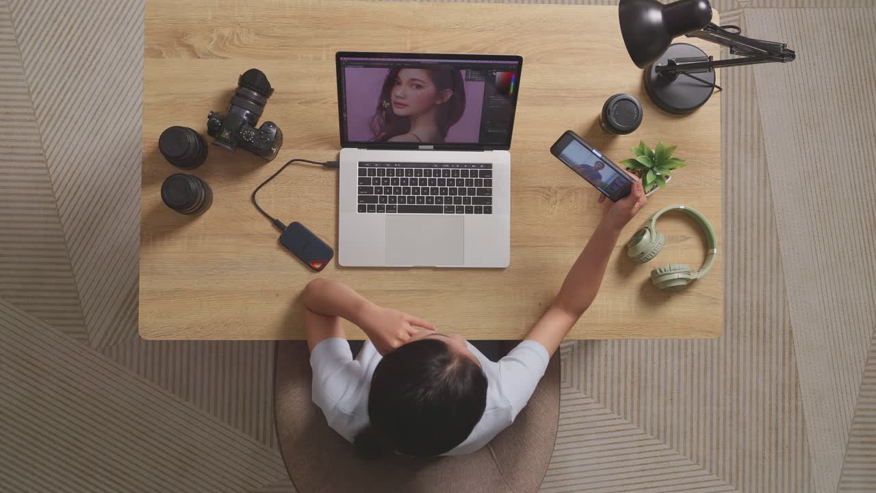 Vista superior de una editora saludando con la mano y haciendo una videollamada en su smartphone mientras está sentada en el espacio de trabajo, usando un portátil junto a la cámara, editando la foto de una mujer en casa.