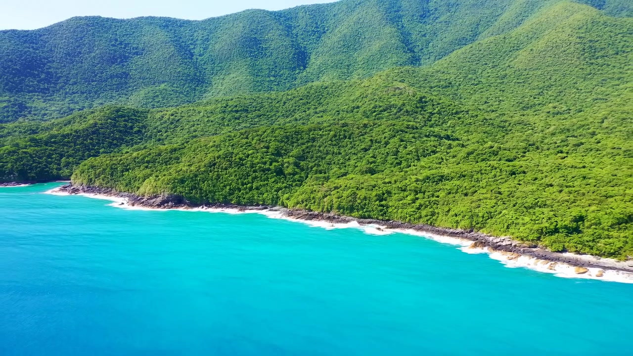 Un punto de vista elevado sigue una costa extensa donde el agua azul verdosa brillante se encuentra con rocas oscuras y espuma, con laderas densamente arboladas que se elevan a través del encuadre bajo una intensa luz solar natural.