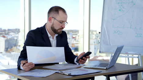 serious businessman in suit working in office. entrepreneur sitting at table using laptop, phone and project papers on window background.