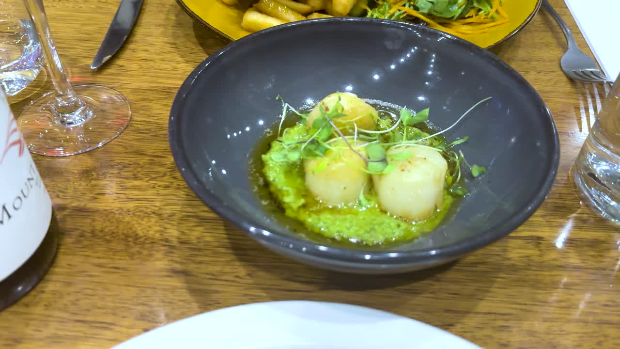 Close-up of a scallop dish with microgreens on a wooden table, surrounded by wine and elegant tableware