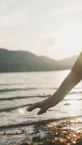 woman dips hand into lake water at sunset closeup. lady touches calm water enjoying beauty of nature in peaceful autumn evening. finding joy in nature