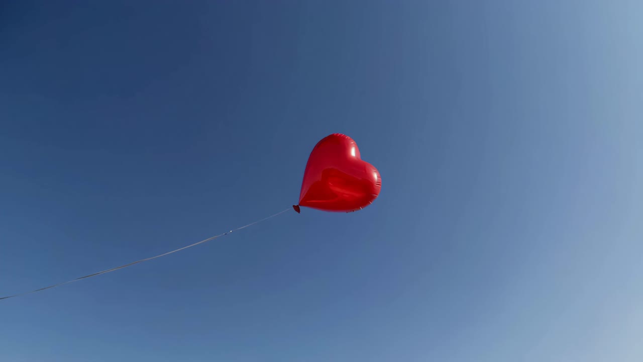 A serene video of a red heart-shaped balloon floating against a clear blue sky, symbolizing love
