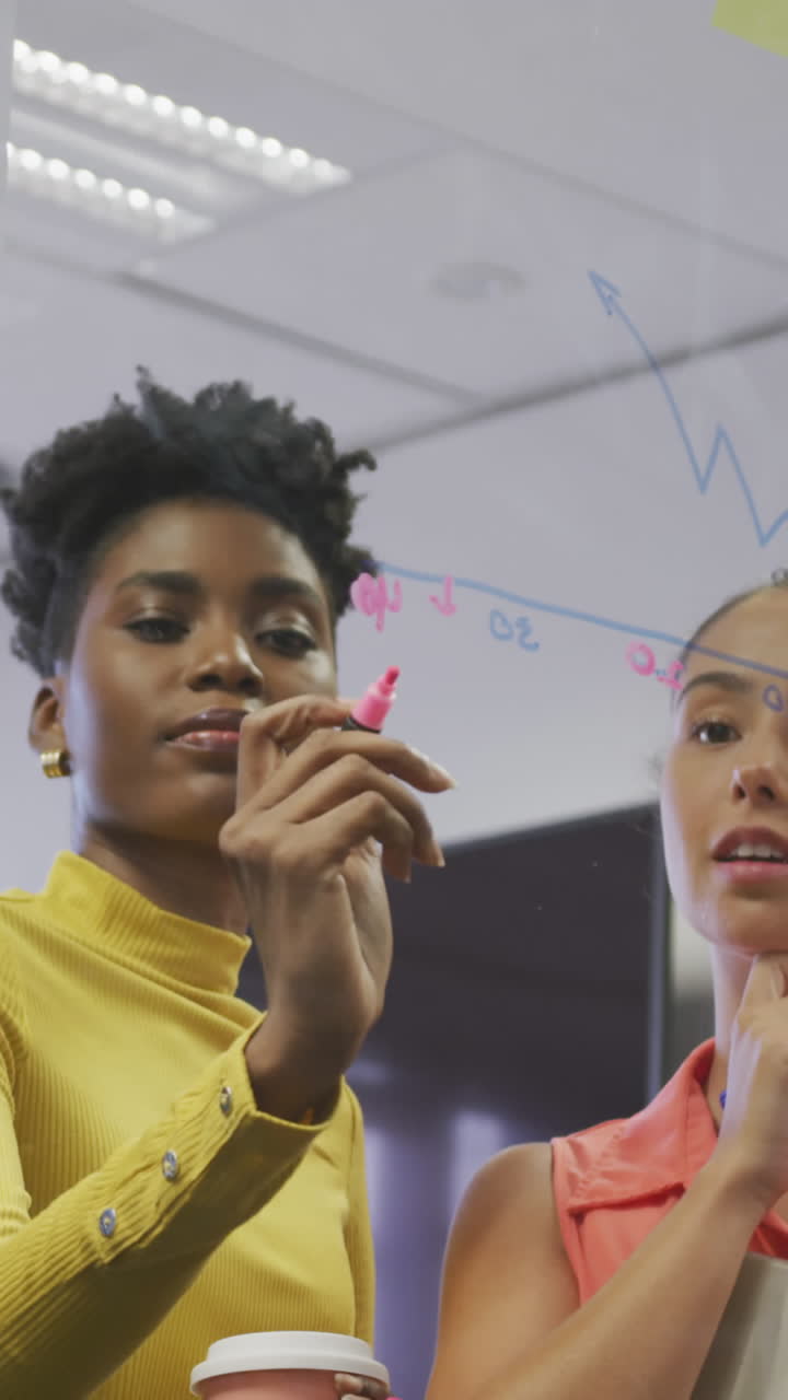 Diverse male and female business colleagues taking notes on glass wall and talking in office