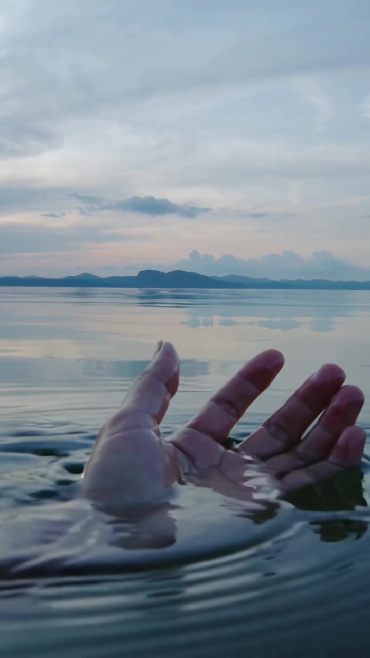 Video verticale: Una mano che entra nell'inquadratura, raccogliendo e sollevando l'acqua, creando increspature e bolle nel lago.