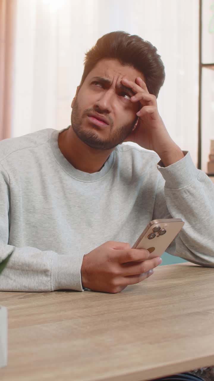 Young indian man texting on smartphone while resting head on hand at table in living room at home