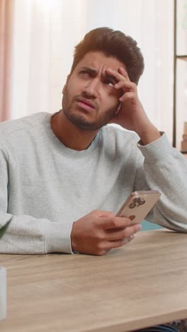 young indian man texting on smartphone while resting head on hand at table in living room at home