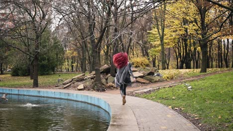 Back-view-of-attractive-woman-with-red-hair-running-towards-pigeons-and-making-them-fly-away-in-a-park-with-artificial-lake