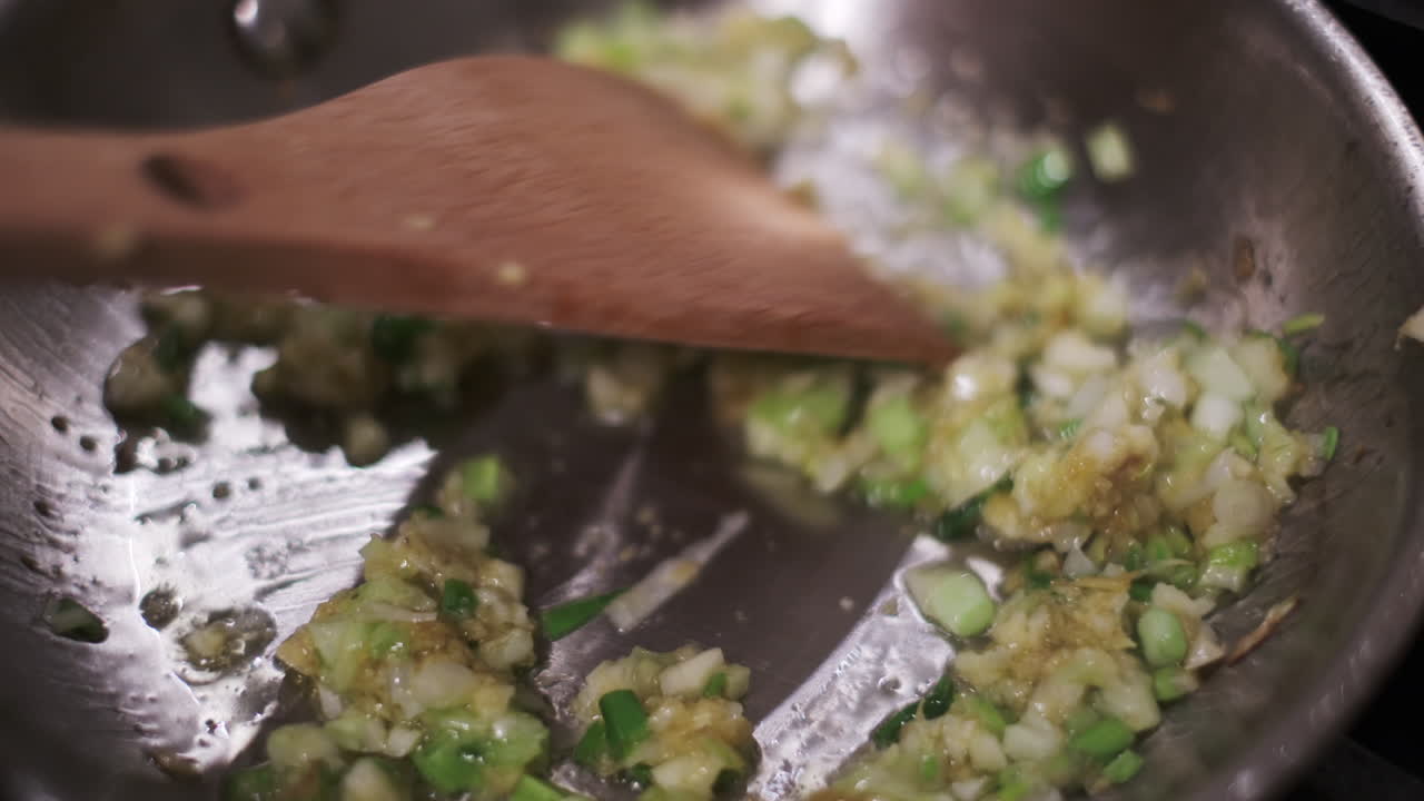 Close-up wooden spatula stirring and turning diced onions, garlic, and shallots as they sizzle and soften
