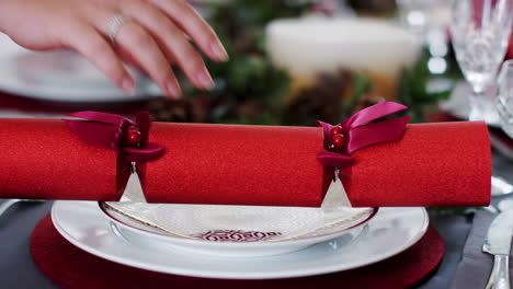 a woman’s hand laying a red christmas cracker on a plate at a table decorated for christmas dinner, close up