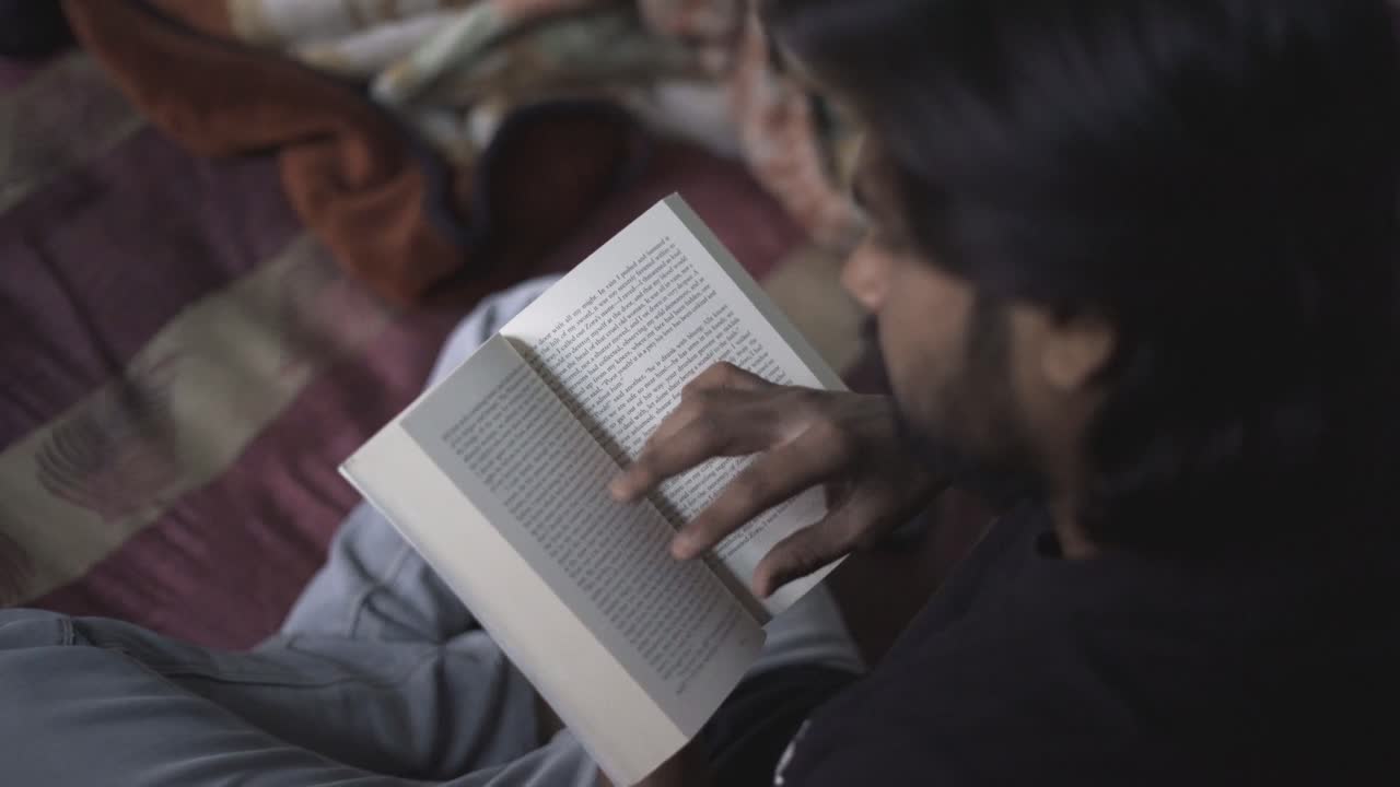 A South Asian Indian reading book in a cold dark room sitting on a bed