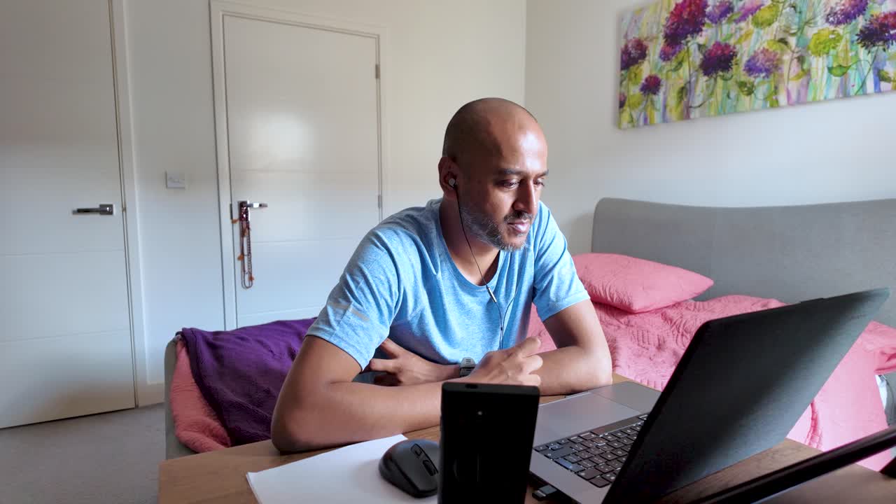 South Asian man working remotely at home, engaged in a video call in bedroom.