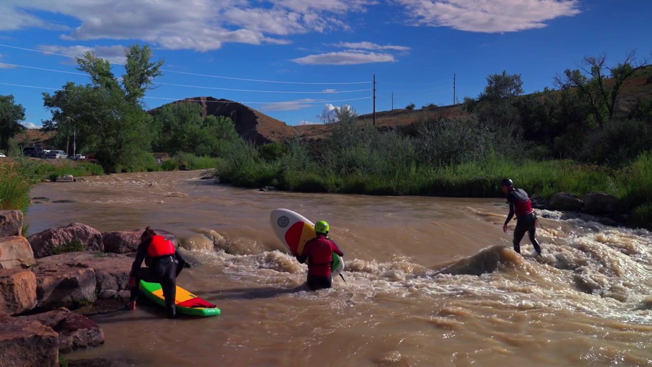 Surfisti fluviali che praticano SUP sulle onde al Montrose Water Sports Park, nella città di Montrose, Colorado. Il paesaggio del fiume Uncompahgre mostra un alto flusso dovuto allo scioglimento della neve e sedimenti fangosi, sotto un cielo blu pomeridiano. Cerise Park, McNeil Fields.