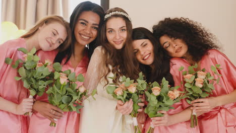 group of multiethnic female friends and bride looking at camera, wearing silk pink and white nighdresses while holding bouquets 2