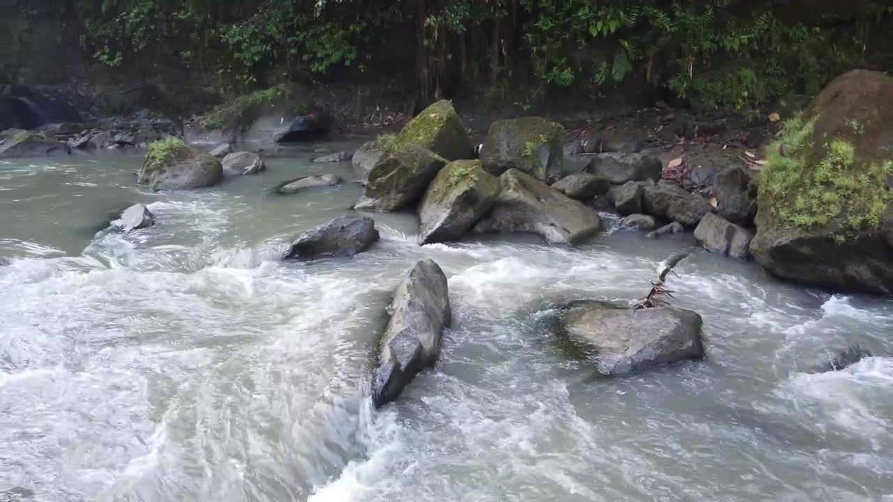 Sungai yang deras di sekitar bebatuan besar di Air Terjun Sumampan di hutan Bali, latar belakang panorama geser kiri