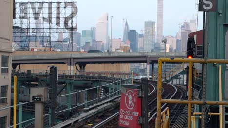 Train-leaving-Queensboro-Plaza-subway-station-on-lower-platform-with-the-Silvercup-Studios-and-the-Manhattan-skyline-in-the-background-filmed-in-the-early-morning-hours-of-a-sunny-day-in-New-York-City