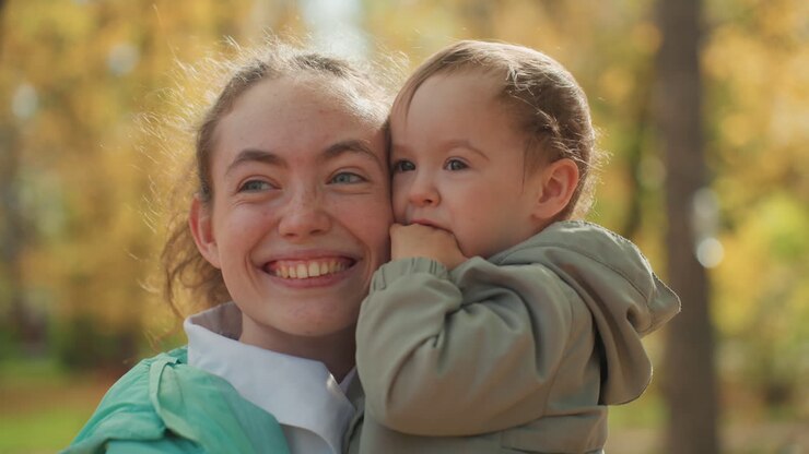 Lächelnde weiße Mutter hält Kleinkind im Park, Herbstsonnenlicht, enge Umarmung, aufrichtiges Porträt, laubreicher Hintergrund, warme Töne, freudige Familienbindung, spielerisches Lachen, sanfter Blickkontakt.