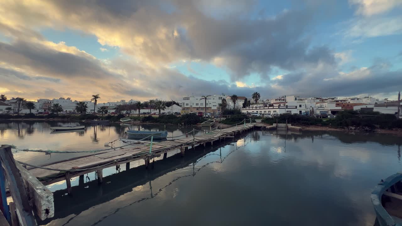 el viejo muelle de madera se extiende a través de aguas tranquilas, que conducen hacia los edificios blancos de la ciudad de barbate bajo un cielo dramático pintado con los cálidos tonos del anochecer en andalucía, españa.