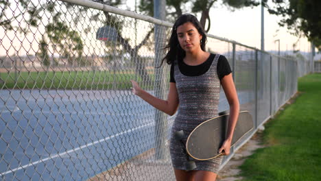 una hermosa joven hispana caminando por un parque urbano con su patineta a lo largo de una cerca de eslabón de cadena a cámara lenta