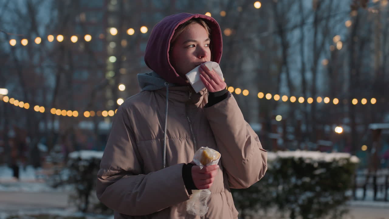 겨울 야외 환경에서 입을 청소하기 위해 킨을 사용하는 동안 한 손으로 간식을 들고 있는 어린 소녀, 배경에 부드러운 보케 조명, 은 날에 근처에서 는 사람들과 함께 음식을 즐기고 있습니다.