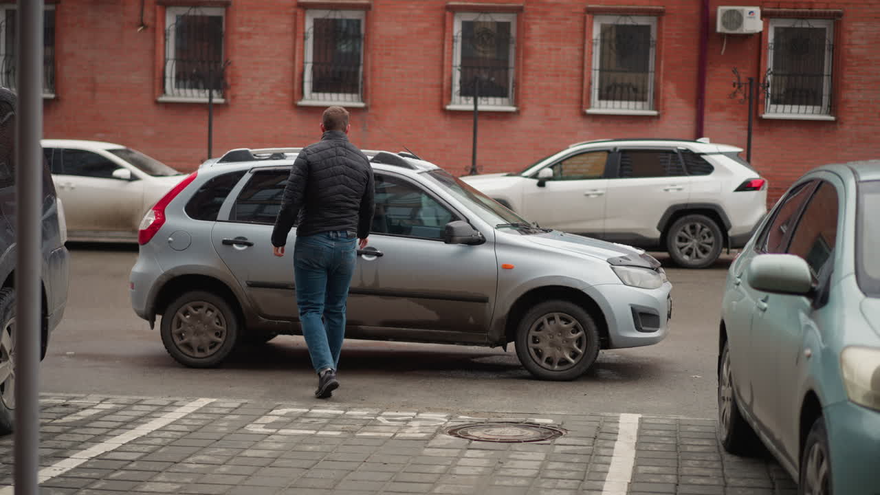 Road traveler walks toward tinted window car parked on urban street, reaches for door handle then opens it and steps inside vehicle for commute, wearing black winter coat and blue jeans