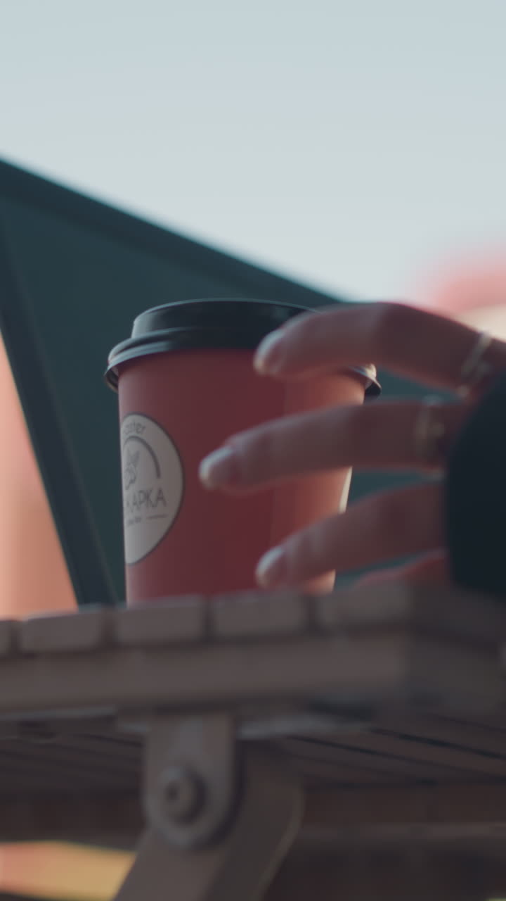 손가락에 반지를 달고 커피 컵을 들고 있는 사람의 클로즈업, 테이블 위에 노트북 에 놓고, 노트북 키보드와 화면이 보이고, 건물의 흐릿한 시야와 밝은 하늘을 배경으로